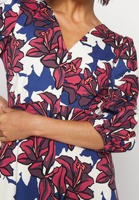 Vestido colorido de longitud hasta la rodilla con un estampado floral en rojo y azul oscuro sobre un fondo blanco, con escote en V y mangas tres cuartos fruncidas.