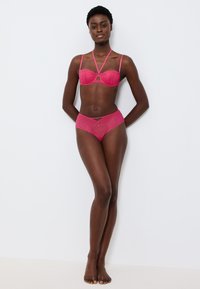 Růžový souprava prádla obsahující podprsenku s geometrickými vzory, podpůrnými kosticemi a odpovídajícími vysokými kalhotkami, oboje se spadlými okraji.