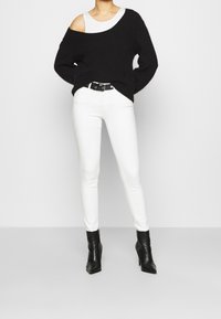 Jersey negro de gran tamaño con un diseño de hombro descubierto, combinado con jeans skinny blancos y botas negras hasta el tobillo. La textura parece suave y de punto.