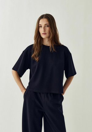 Jonge vrouw met lang bruin haar, gekleed in een losse zwarte T-shirt en bijpassende zwarte broek, staand met handen in de zakken tegen een effen achtergrond.