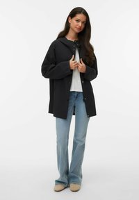 Veste noire à capuche avec boutons-pression, portée sur un haut blanc. Associée à un jean évasé bleu clair et des chaussures beige. Texture lisse.