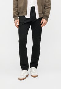 Μαύρο παντελόνι με ίσια κοπή και λεία υφή, συνδυασμένο με λευκά αθλητικά παπούτσια και χακί φούτερ με φερμουάρ.