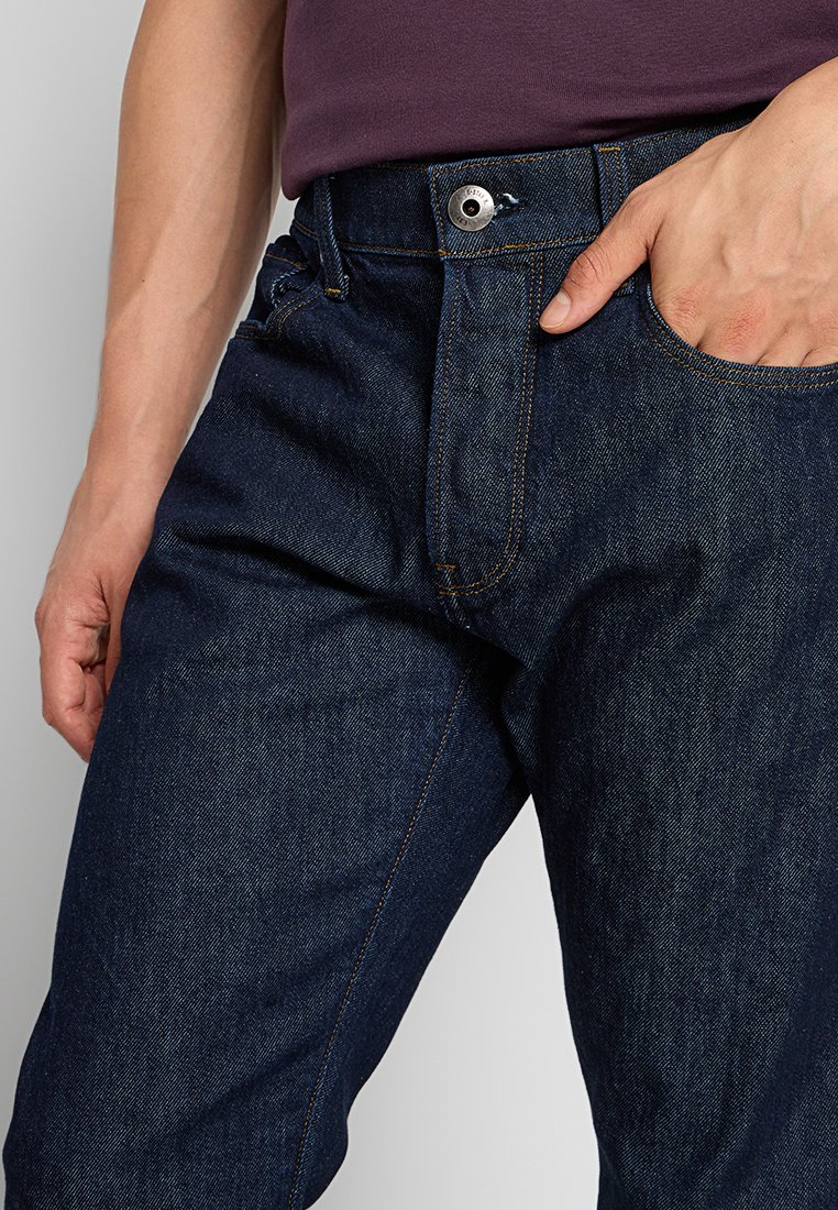 Uomo che indossa jeans di denim blu scuro e una maglietta viola, con la mano destra in tasca e la mano sinistra rilassata lungo il fianco.