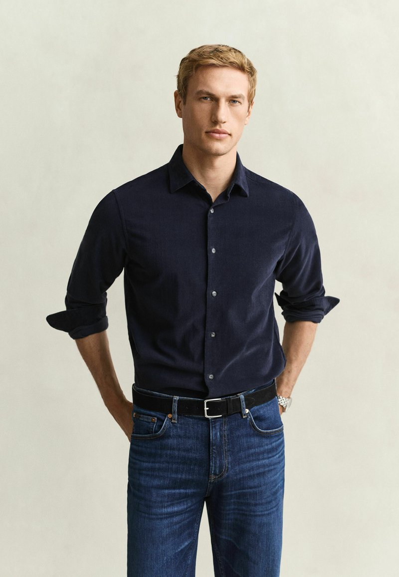 Blond man wearing a navy button-up shirt with rolled sleeves and blue jeans, standing with hands behind his back against a plain background.