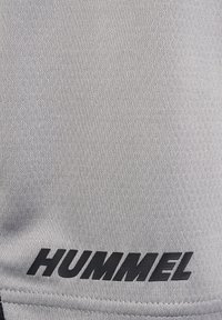 Grå atletisk tyg med en slät textur; har en svart "HUMMEL"-logotyp tryckt tydligt. Söm Detalj syns längs kanten.