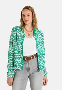 Chaqueta bomber con patrón verde y crema que presenta una textura suave, cuello acanalado y puños elásticos, llevada sobre una blusa blanca y jeans grises.