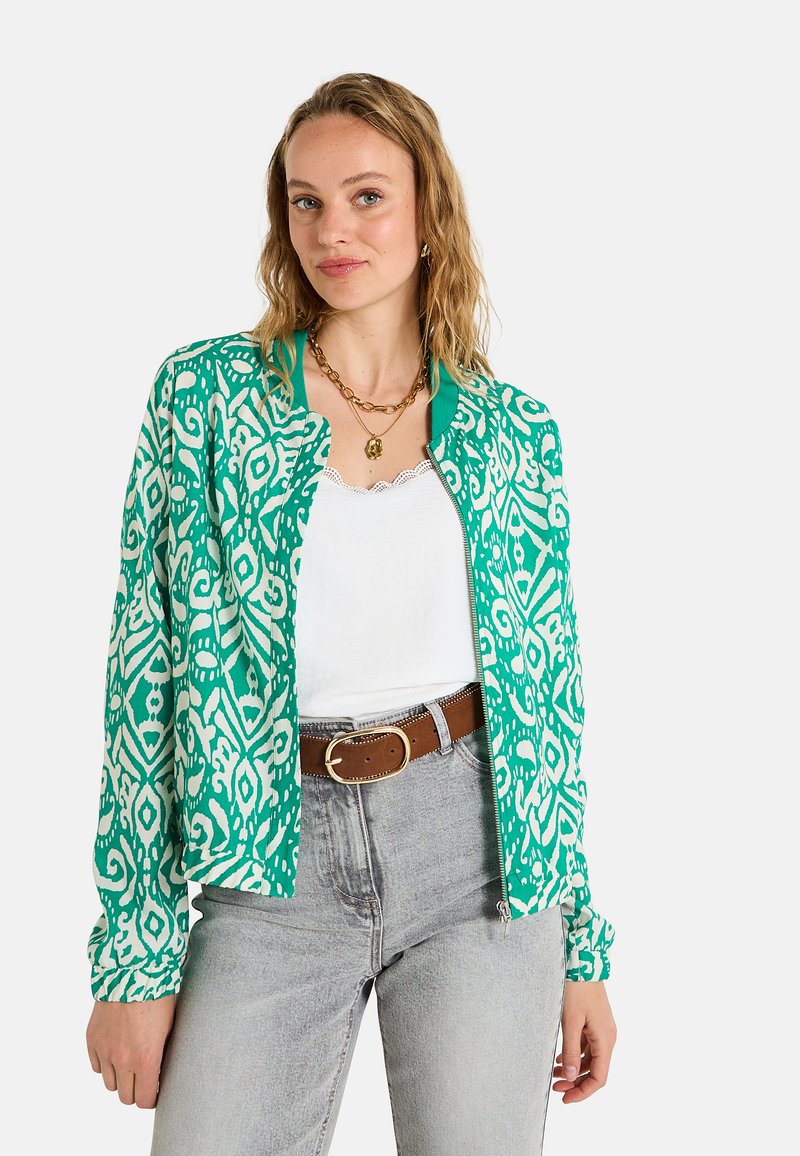 Chaqueta bomber con patrón verde y crema que presenta una textura suave, cuello acanalado y puños elásticos, llevada sobre una blusa blanca y jeans grises.