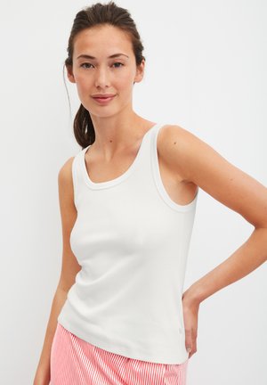 Jonge vrouw met donker haar in een staart, gekleed in een wit mouwloos geribd topje en rood-wit gestreepte broekjes, staand tegen een effen achtergrond.