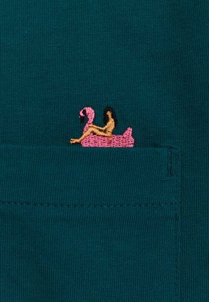 Teal stof heeft een kleine geborduurde roze flamingo op een gele drijver met bruine accenten, gepositioneerd in een zakgebied.