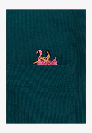 Teal stof heeft een kleine geborduurde roze flamingo op een gele drijver met bruine accenten, gepositioneerd in een zakgebied.