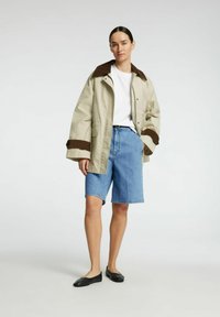Beige jacka med bruna detaljer, vit skjorta, blå denim shorts och svarta flator. Har stor krage och avslappnad passform.