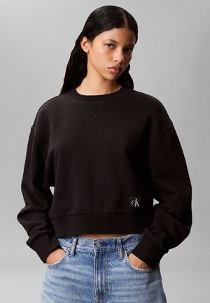 Giovane donna con capelli lunghi e scuri indossa una felpa nera Calvin Klein e jeans azzurri, con le mani infilate nelle tasche dei jeans, sfondo neutro.