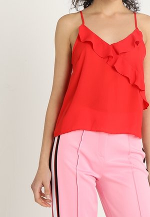 Rode mouwloze top met een geplooid paneel bij de halslijn, gecombineerd met lichtroze broek met zwarte strepen aan de zijkant. Gladde stoftextuur.