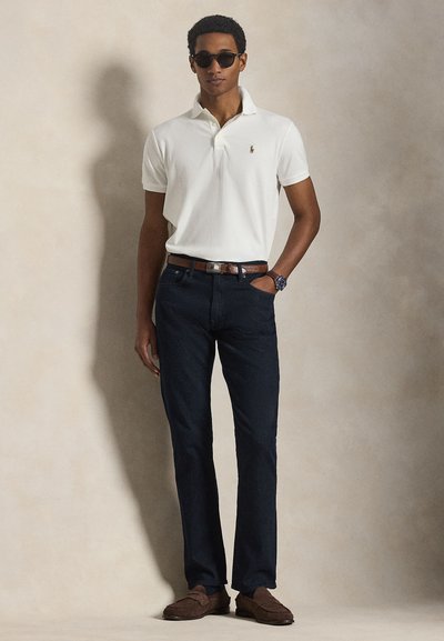 Férfi fehér rövid ujjú pólóban, sötét farmerben, barna övvel és cipővel, napszemüvegben és karórával, semleges háttér előtt állva.