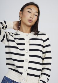 Una giovane donna con lunghi capelli scuri indossa orecchini a cerchio d'oro e un cardigan crema con strisce nere orizzontali e grandi bottoni rotondi.