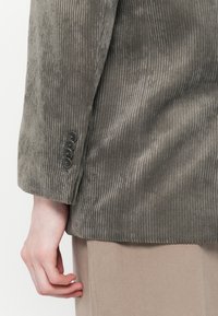 Gray corduroy blazer sleeve with ribbed texture, featuring four black buttons at the cuff. Beige fabric underneath visible at the hem.