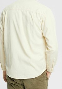 Camicia a maniche lunghe giallo chiaro con dettaglio a yoke sul retro. Presenta polsini con bottone e un orlo curvo. Tessuto morbido, simile al cotone.