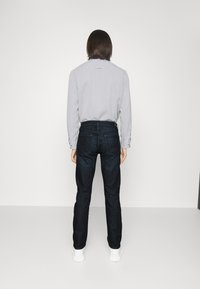 Cămașă lungă gri deschis cu detalii cu nasturi, asociată cu blugi dark blue slim-fit. Modelul poartă adidași albi, văzut din spate.
