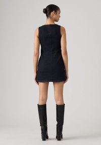 Γυναίκα με κότσο στα μαλλιά, φορώντας ένα μαύρο μίνι φόρεμα από τζιν χωρίς μανίκια και μαύρες μπότες μέχρι το γόνατο με τακούνια, στέκεται μπροστά σε ένα απλό φόντο.