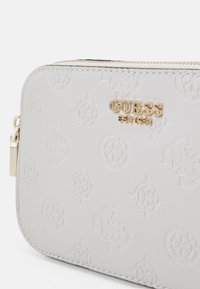 Biała tłoczona syntetyczna skórzana saszetka z złotym logo "GUESS", z teksturowanym wzorem i zapięciem na zamek.