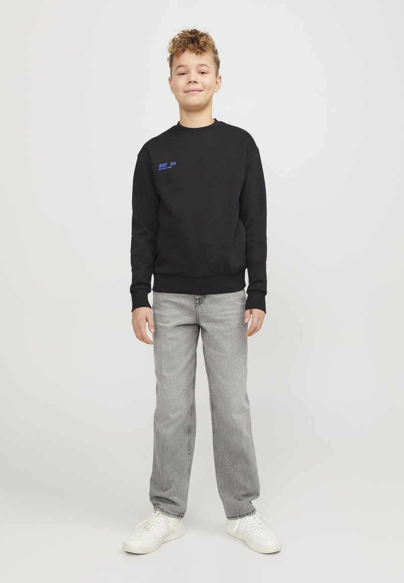 Felpa nera con testo blu, jeans grigi e sneaker bianche. Design aderente con polsini e vita a costine. Modello in piedi contro uno sfondo neutro.