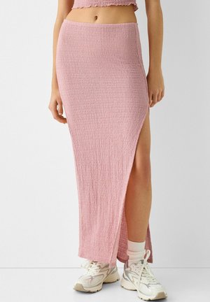 Vrouw die een roze midi-rok met textuur en een zijsplit draagt, witte sokken en witte sportsneakers, staand tegen een effen achtergrond.