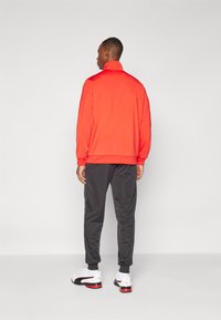Rode polyester jas met hoge kraag, gecombineerd met grijze sweatpants en witte sportschoenen met zwarte en rode accenten.