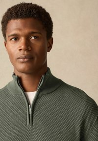 Grüner strukturierter halber Reißverschluss-Pullover mit hohem Kragen und gerippten Kanten, getragen über einem weißen Hemd mit dezentem Wabenmuster.