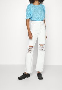 Blusa floreale azzurro chiaro con maniche a sbuffo, abbinata a jeans bianchi a vita alta con grandi strappi sulle ginocchia e scarpe slip-on spesse nere.