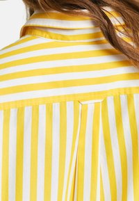 Camicia con colletto caratterizzata da strisce verticali gialle e bianche. Tessuto liscio con un leggero lucido e dettaglio plissettato sul retro.