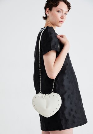 Heart-shaped white bag with silver studs, smooth texture, and chain strap. Worn over a black textured dress, showcasing its unique design.