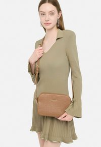 Borsa a tracolla marrone con logo in rilievo, caratterizzata da una texture liscia e una tracolla regolabile. Indossata con un abito aderente a coste verde oliva.
