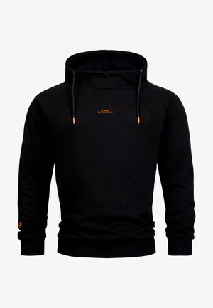 Smilende mand iført en sort hættetrøje med teksten "ICONIC EXPERIENCE" og blå jeans, stående mod en ensfarvet hvid baggrund.