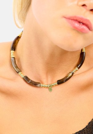 Collier composé de segments en résine brun foncé avec des accents de perles dorées et un petit charme en or, reposant sur une clavicule nue.