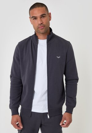 Man wearing a dark gray zip-up jacket over a white t-shirt, standing against a plain light background.