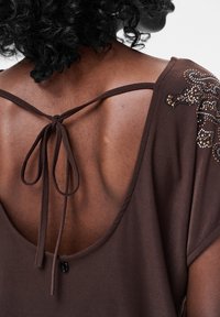 Brązowy top z szerokim dekoltem na plecach, z zapięciem na wiązaną tasiemkę oraz ozdobami z kamieni szlachetnych na jednym ramieniu. Miękka faktura.