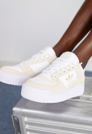 Witte en beige Adidas sneakers met logo aan de zijkant, gedragen door donkergekleurde voeten die rusten op een zilverkleurige metalen koffer.