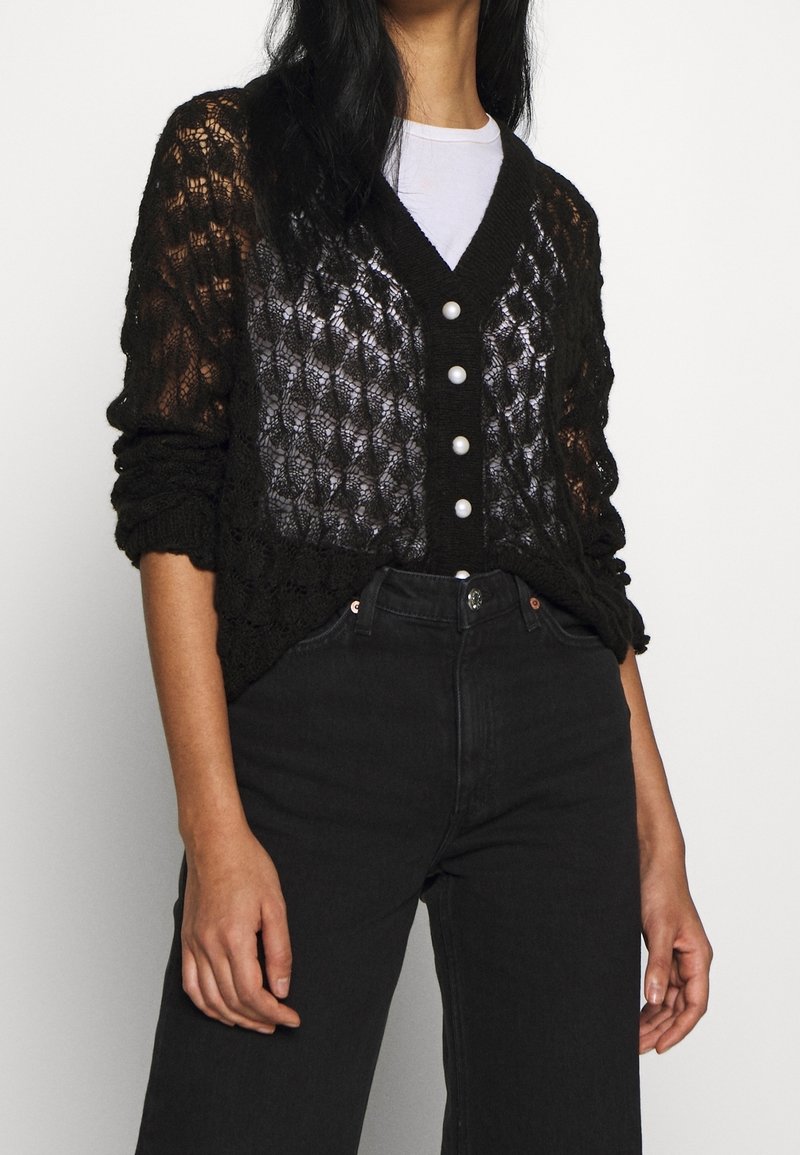 Kvinna klädd i en svart spetsstickad kavaj med pärlknappar över en vit skjorta, kombinerat med högmidjade svarta jeans.