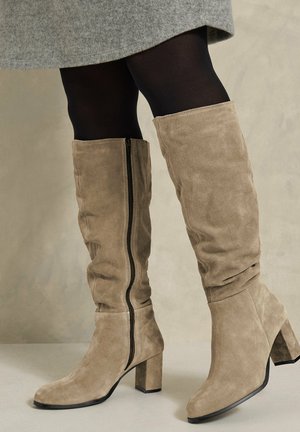 Femme portant des bottes hautes taupe en daim avec talons blocs, des collants noirs et une jupe grise sur un fond neutre.