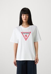 Bílý bavlněný tričko s krátkým rukávem, které má uprostřed červené trojúhelníkové logo se slovem "GUESS" a otazníkem.
