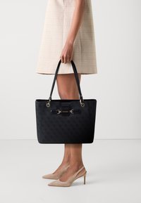 Sac fourre-tout noir avec un design texturé, des accents en métal doré, des poignées doubles et une forme rectangulaire. Le nom de la marque "GUESS" est affiché de manière proéminente.