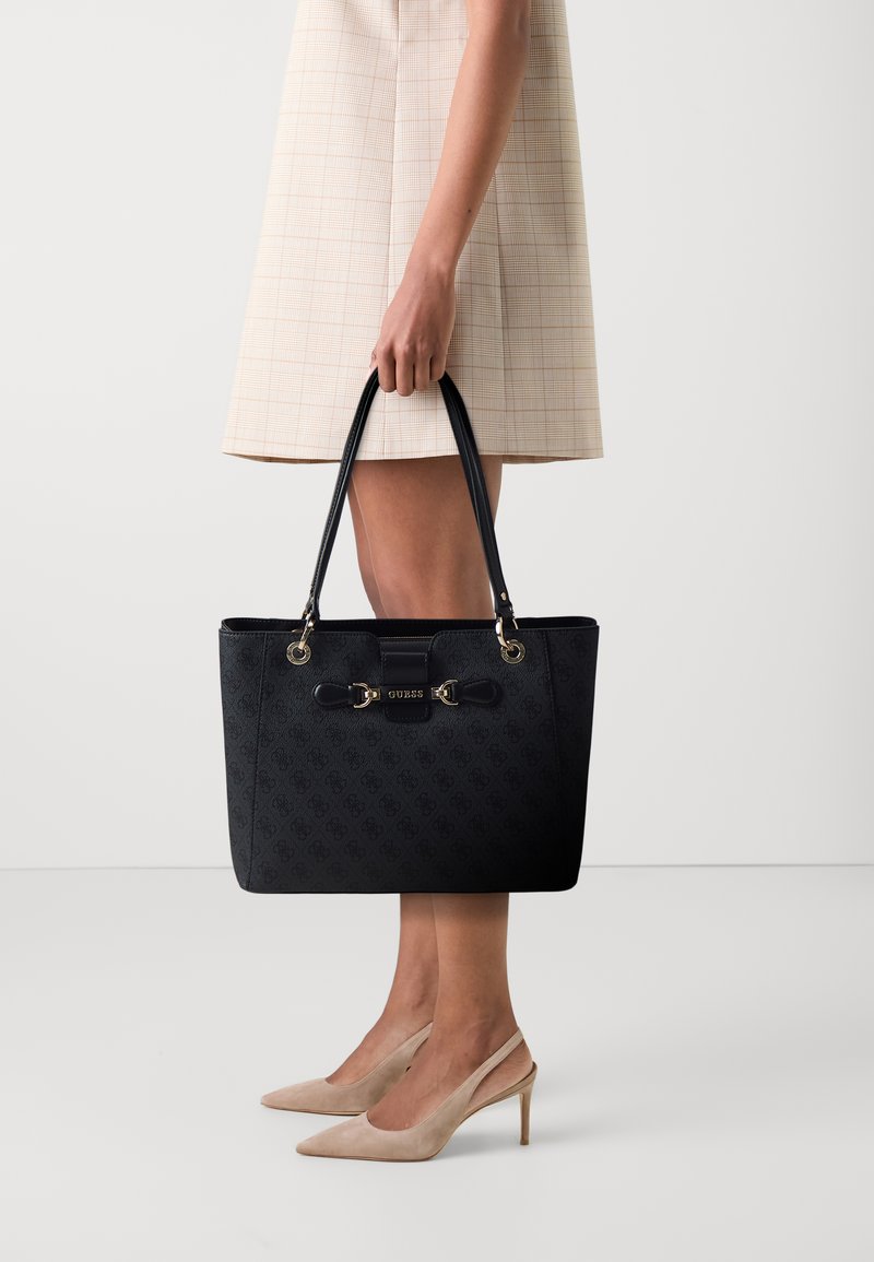 Sac fourre-tout noir avec un design texturé, des accents en métal doré, des poignées doubles et une forme rectangulaire. Le nom de la marque "GUESS" est affiché de manière proéminente.