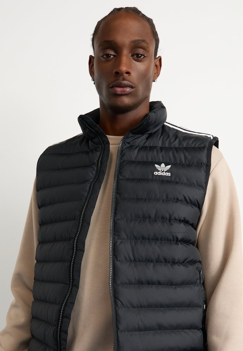 Czarna kamizelka puchowa z wysokim kołnierzem, zamkiem błyskawicznym z przodu i subtelnym poziomym przeszyciem. Logo Adidas po lewej stronie klatki piersiowej. Miękka faktura.