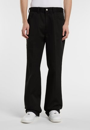 Mies, jolla on päällään väljät mustat housut, valkoiset tennarit ja valkoinen hihaton paita, seisoo yksinkertaisella vaalealla taustalla.