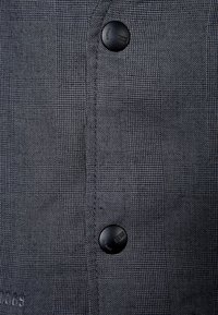 Šedá kostkovaná látka košile s texturovaným proplétáním, obsahující tři černé plastové knoflíky a viditelné švy podél légů.