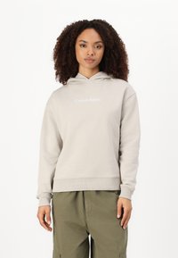 Sivá mikina s kapucňou na šnúrku a rebrovanými manžetami, s bielym logom "Calvin Klein". Skombinovaná so zelenými cargo nohavicami.