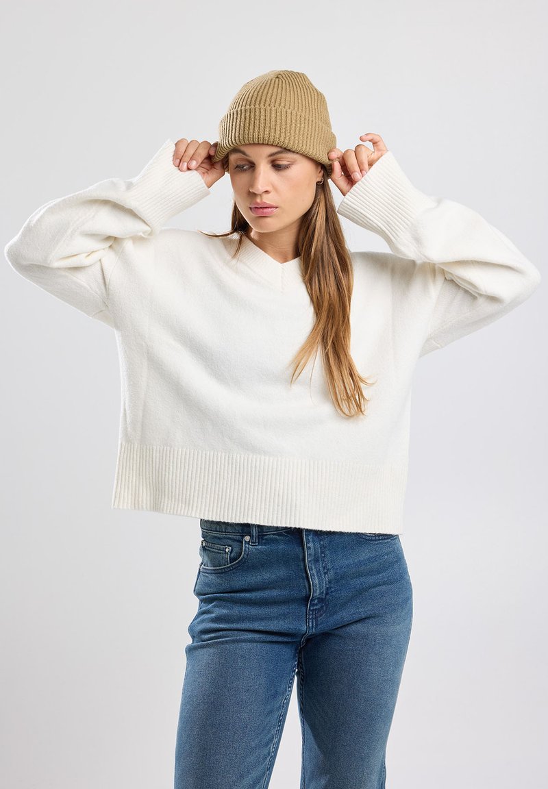 Pull en V coupé blanc, avec des poignets et un ourlet côtelés. Associé à un jean bleu ; mannequin portant un bonnet en tricot beige. Design simple et moderne.