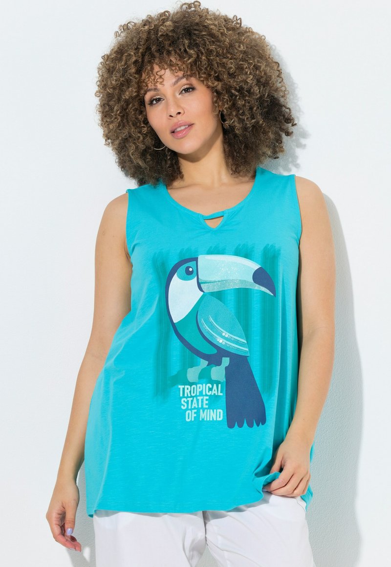 Donna con capelli ricci indossa un top turchese senza maniche con una grande grafica di tucano e la scritta "Tropical State of Mind", abbinato a pantaloni bianchi.