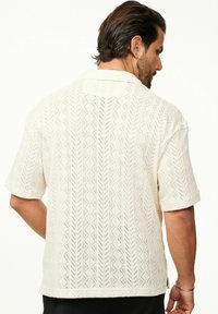 Hellbeige Hemd mit kurzen Ärmeln, das ein strukturiertes Strickmuster mit Zickzack-Designs und einem flachen Kragen aufweist. Atmungsaktives Material, lockere Passform.
