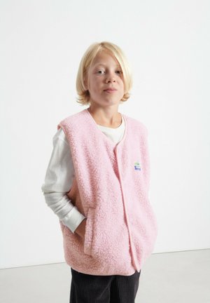Fleece vest in light pink with front pockets and a button closure, worn over a white long-sleeve shirt, featuring a small tag detail.